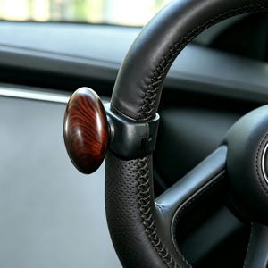 Puede incluir: Un pomo para volante con un mango ovalado de acabado en veta de madera marrón oscuro pulido, unido a un soporte de montaje negro. El pomo está asegurado a un volante forrado en cuero negro con costuras visibles y textura perforada.