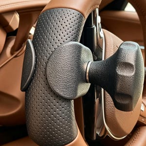 May include: A black steering wheel spinner knob with a textured grip surface, attached to a perforated black leather steering wheel cover. The knob features a contoured, ergonomic shape and is mounted on a brown leather steering wheel.