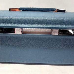 May include: A rectangular blue hard-shell vanity case with a textured exterior finish. The case features a top handle, a silver-toned metal trim around the center, and two silver-toned latches on the front side for secure closure.