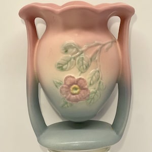 May include: A ceramic vase featuring a scalloped rim and two side handles. The body displays a gradient color transition from soft pink at the top to a muted blue at the base. A raised relief of a pink flower with green leaves is centered on the front.