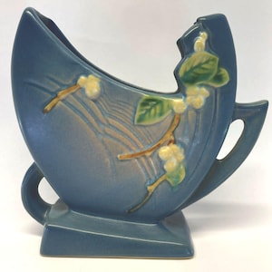May include: A matte blue ceramic vase with a curved, asymmetrical silhouette and a rectangular base. The surface features a raised, molded branch design with green leaves and pale yellow berries, accented by subtle tan-colored linear detailing.