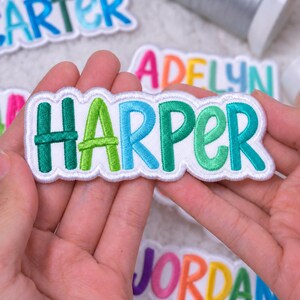 以下が含まれることがあります： 緑、ライム、青のグラデーションの糸で「HARPER」という名前が刺繍されたカスタムワッペン。文字は太く丸みを帯びたフォントで縫い付けられており、白い背景に白いスカラップ状の縁取りが施されています。