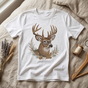 May include: A white short-sleeved t-shirt featuring a detailed graphic of a brown deer head with large antlers. The deer is surrounded by a natural arrangement of pine needles, pinecones, wheat stalks, and oak leaves in earthy brown and green tones.