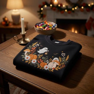 May include: A black crewneck sweatshirt featuring an embroidered design of two white ghosts, orange pumpkins, mushrooms, white daisies, and autumn foliage. The embroidery is arranged in a curved pattern across the chest of the garment.