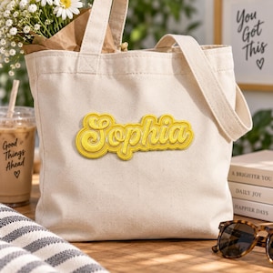 Peut inclure: Un sac fourre-tout en toile de couleur crème avec un écusson texturé jaune de style rétro portant le prénom Sophia en écriture cursive au centre. Le sac est doté de deux anses en tissu robustes et d'une ouverture simple sur le dessus.