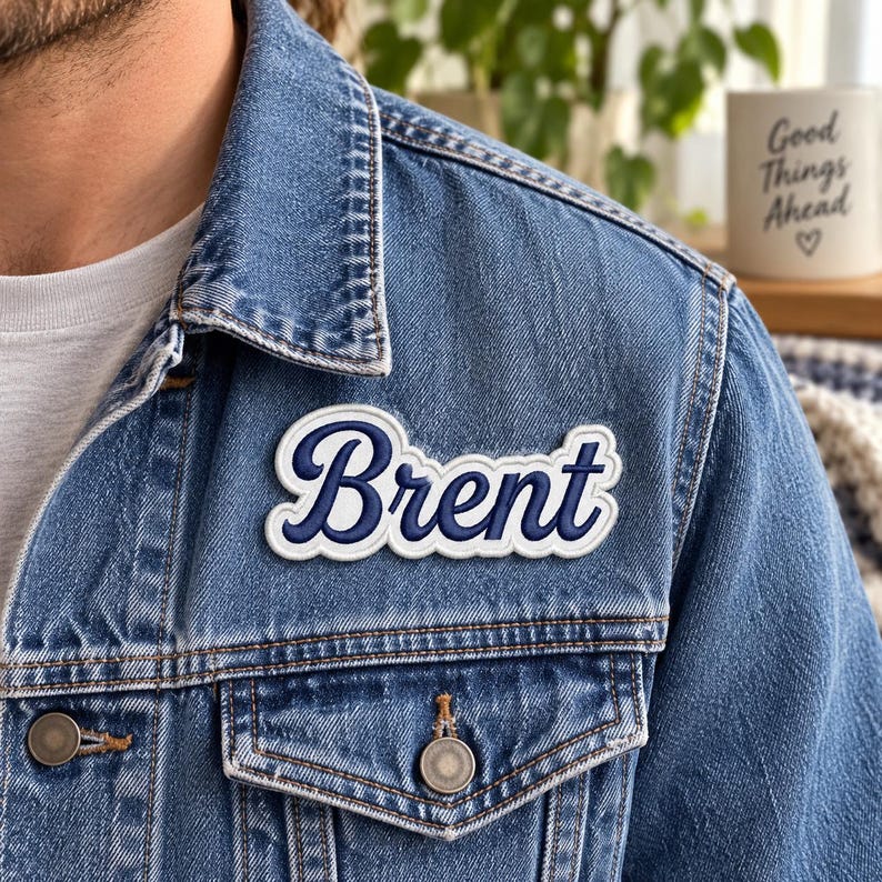 以下が含まれることがあります： 筆記体でBrentという名前が刺繍された、白と濃紺のワッペンが付いた青いデニムジャケット。ワッペンはジャケットの胸ポケットの上に配置されており、無地の白いTシャツの上に着用されています。