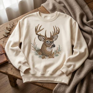 May include: A cream-colored crewneck sweatshirt featuring a detailed embroidered graphic of a brown deer with large antlers, surrounded by pine needles, pinecones, and dried leaves centered on the front of the garment.