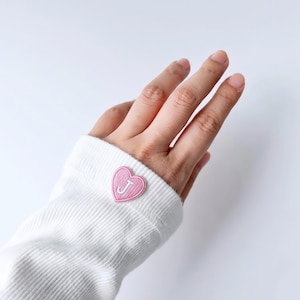 Op de afbeelding: Een wit shirt met lange mouwen van geribbelde stof, voorzien van een kleine, roze geborduurde hartvormige patch op de manchet. Het hart bevat de witte hoofdletter J in het midden, wat zorgt voor een duidelijk detail op de mouw.