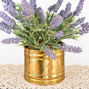 May include: A bouquet of artificial purple lavender flowers with green stems and leaves arranged in a cylindrical, polished gold-colored metal pot. The container features horizontal ridges around the top and bottom edges with a reflective surface.