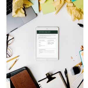 May include: A white tablet displaying a digital journal titled The Mind-Body Compass Journal with a dark green header. The screen shows a daily blueprint template with structured sections for goals, tasks, and checklists, presented on a white surface.
