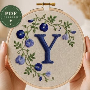 May include: A circular wooden embroidery hoop containing a beige fabric panel with a hand-stitched letter Y in dark blue thread, surrounded by a delicate vine of green leaves and blue butterfly pea flowers. A green circular PDF PATTERN badge.