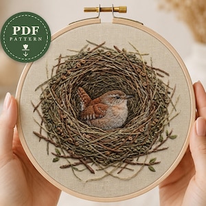 May include: A circular wooden embroidery hoop containing a fabric panel with a detailed needlepoint design of a small brown bird sitting inside a textured nest made of brown and green thread, with a green circular PDF pattern icon in the corner.