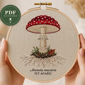Può includere: Un telaio da ricamo circolare in legno contenente un pannello in tessuto con un ricamo dettagliato di un fungo Amanita muscaria rosso e bianco. Il disegno presenta radici visibili, muschio verde e il testo Amanita muscaria FLY AGARIC.