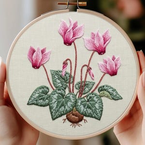 May include: A circular wooden embroidery hoop containing a fabric panel with a hand-stitched design of a cyclamen plant. The embroidery features vibrant pink flowers, dark green heart-shaped leaves with light green veining, and a brown bulb.