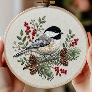 May include: A circular wooden embroidery hoop containing a fabric panel with a detailed needlepoint design of a black-capped chickadee perched on a pine branch. The scene includes brown pinecones, green pine needles, and clusters of red berries.
