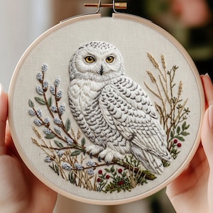 以下が含まれることがあります： 枝にとまった白いシロフクロウの精巧な刺繍デザインが施された円形の木製刺繍枠。フクロウは黄色い目と灰色の羽のディテールが特徴で、青い蕾と赤い実の刺繍が施された小枝に囲まれています。明るい色の布地に刺繍が施されています。