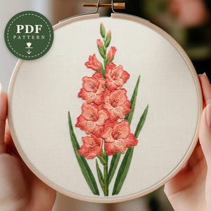 May include: A circular wooden embroidery hoop containing white fabric with a hand-stitched gladiolus flower in shades of coral and pink, featuring green leaves and stems. A green circular badge in the top left corner displays the text PDF PATTERN.