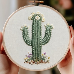 May include: A circular wooden embroidery hoop containing a fabric panel with a hand-stitched green cactus design. The cactus features white flowers at the top and on its two arms, with small white dots representing spines and colorful floral accents.