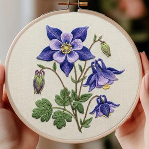 May include: A circular wooden embroidery hoop containing a fabric panel with a detailed needlepoint design of purple and lavender columbine flowers, green leaves, and small buds, featuring a bright yellow center on the primary bloom.