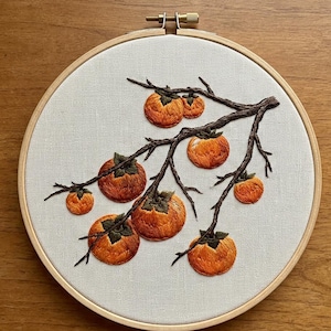 May include: A circular wooden embroidery hoop containing white fabric with a hand-stitched design of a brown tree branch bearing multiple orange persimmon fruits. The embroidery features detailed thread work in shades of orange and brown.