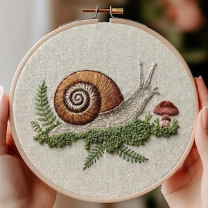 Könnte beinhalten: Ein runder Stickrahmen aus Holz mit einem cremefarbenen Stoff. Der Stoff zeigt ein handgesticktes Motiv einer braunen Schnecke mit spiralförmigem Gehäuse, die auf grünem Moos ruht, umgeben von grünen Farnblättern und einem kleinen braunen Pilz.