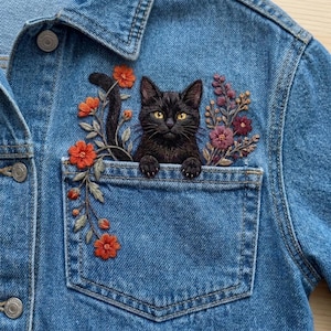 Op de afbeelding: Een blauw spijkerjack met een borduursel van een zwarte kat met gele ogen die uit de borstzak kijkt. Het borduurwerk bevat oranje en rode bloemen aan de linkerkant en paarse en roze bloemtakjes aan de rechterkant van de borstzak.