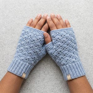 May include: A pair of light blue fingerless gloves featuring a textured shell stitch pattern throughout the main body. The gloves have a ribbed cuff at the wrist and a small, rectangular tan leather tag attached to the bottom edge of each piece.