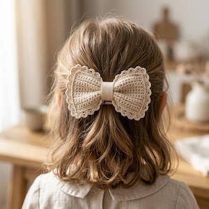 Può includere: Un fiocco per capelli all'uncinetto color beige con bordo smerlato e una fascia centrale, fissato tra i capelli castano chiaro. Il fiocco presenta un motivo a punti denso e strutturato con un bordo decorativo che ne definisce la forma.