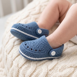 May include: A pair of blue crochet baby shoes featuring a textured knit pattern, a white horizontal stripe along the sole, and a white button detail on the side strap. The shoes have small ventilation holes across the top of the toe area.