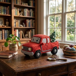 Op de afbeelding: Een rood gehaakt speelgoedpickup met grijze accenten en een groene gehaakte kerstboom in de laadbak. De truck heeft witte en zwarte ogen op de voorruit, een grijze voorbumper en zwarte wielen, geplaatst op een houten ondergrond.