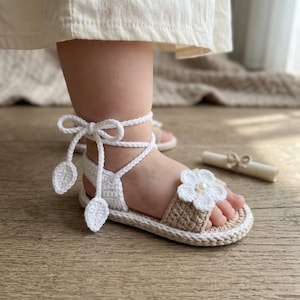 May include: A pair of handmade crochet baby sandals featuring a tan base, a white crocheted flower with a central pearl-like bead on the toe strap, and white braided ankle ties that wrap around the leg, finished with small crocheted leaf accents.