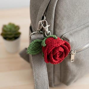 May include: A red crocheted rose keychain with a green leaf attached to a silver metal lobster clasp. The floral accessory is clipped onto the zipper pull of a grey canvas backpack, featuring detailed yarn stitching and layered petals.