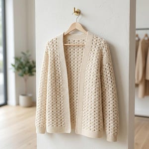 Peut inclure: Un gilet en crochet de couleur crème, ouvert sur le devant, présentant un motif de points en éventail répété sur tout le corps et les manches, avec une bordure côtelée unie le long des bords avant, de l'encolure et des poignets, sur un cintre.