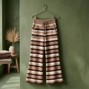 Puede incluir: Pantalones de ganchillo con un patrón de rayas horizontales en tonos marrón oscuro, marrón claro y crema. Los pantalones tienen una silueta de pierna ancha, una cintura elástica acanalada con un cordón a juego y cuelgan de una percha.
