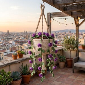 May include: A cream-colored crocheted hanging planter featuring long, trailing vines with small purple crocheted roses and green leaves. The planter is suspended by a braided cord and includes a decorative tassel at the bottom center.