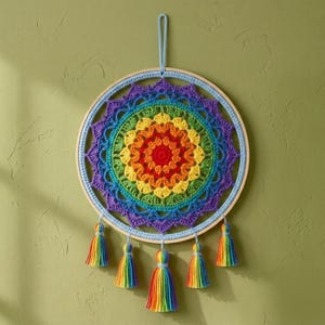May include: A circular wall hanging featuring a vibrant, multi-colored crochet mandala centered within a light-colored wooden hoop. The design displays concentric rings of red, orange, yellow, green, blue, and purple yarn, finished with five hanging tassels in matching rainbow colors and a light blue yarn loop for suspension.