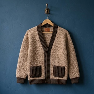 Könnte beinhalten: Eine beige gehäkelte Strickjacke mit einem dunkelbraunen gerippten V-Ausschnitt, passenden dunkelbraunen gerippten Bündchen, einem dunkelbraunen gerippten Saum und zwei dunkelbraunen gerippten aufgesetzten Taschen auf der Vorderseite.