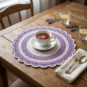 May include: A round, crocheted placemat featuring concentric rings of white and lavender yarn with a scalloped edge. A white ceramic teacup and saucer containing tea and a single purple flower petal sit centered on the placemat.