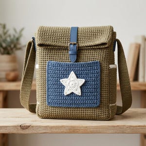 May include: An olive green crochet crossbody bag featuring a blue rectangular front pocket with a white crocheted star appliqué. The bag has a fold-over flap secured by a blue faux leather strap with a metal buckle and a wide crochet shoulder strap.