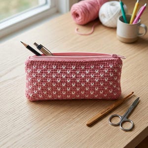 Può includere: Un astuccio rettangolare all'uncinetto color rosa polvere con un motivo ripetuto a forma di V bianca. L'astuccio presenta una chiusura a zip rosa abbinata sul bordo superiore e contiene una matita nera e una penna argentata.