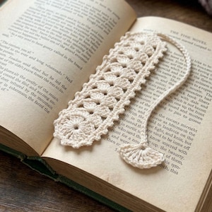 Op de afbeelding: Een gebroken witte gehaakte boekenlegger met een herhalend cirkelvormig bloemmotief en een geschulpte rand. Een dun gehaakt koord loopt vanaf de bovenkant en eindigt in een kleine, halfronde waaiervormige kwast op de open bladzijde.