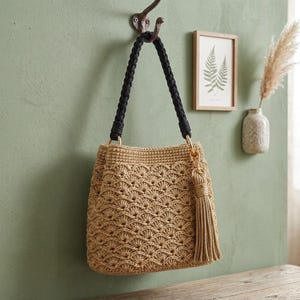 May include: A beige crochet handbag featuring a textured fan-shaped stitch pattern. The bag includes a thick black braided handle attached with gold-toned metal rings and a matching beige tassel accessory hanging from one side of the bag's top edge.