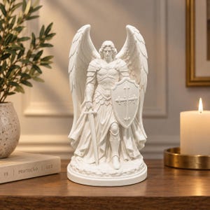 Op de afbeelding: Een wit mat afgewerkt beeldje van een aartsengel op een ronde voet. De figuur draagt een gedetailleerd harnas, houdt een lang zwaard in de rechterhand en een schild met een kruisembleem in de linkerhand, voorzien van grote vleugels.