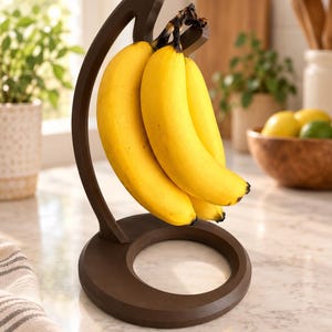 Peut inclure: Un porte-bananes marron foncé doté d'un bras incurvé et d'une base circulaire. Le support présente un crochet sur le dessus pour suspendre un régime de bananes jaunes. La structure est faite d'un matériau solide avec une finition lisse.