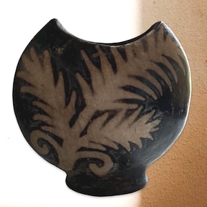 May include: A rounded ceramic vase with a concave top edge, featuring a matte black finish and a contrasting cream-colored fern or leaf pattern painted on the front. The vessel has a narrow base and a wide, curved body with a smooth texture.