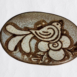 May include: An oval ceramic dish featuring a speckled light blue and brown glaze. The surface displays a raised, embossed design of a bird perched atop a large, swirling flower, with the bird and flower rendered in a textured, creamy off-white finish.