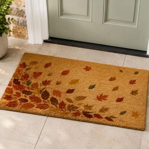 May include: A rectangular coir doormat featuring a natural tan base color with a printed design of scattered autumn leaves in shades of red, orange, brown, and dark green. The leaves are concentrated on the left side and drift across the mat.