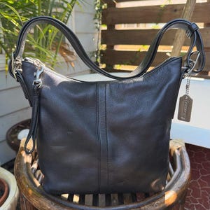 May include: A black leather hobo bag featuring a single shoulder strap, a top zipper closure, and a silver-toned metal hangtag attached to the side. The bag has a vertical center seam and silver-toned hardware, including a clip on the strap.