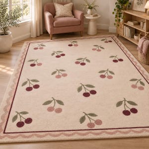 May include: A rectangular area rug with a cream-colored background featuring a repeating pattern of red and pink cherries with green stems. The rug is framed by a dark red border and an outer scalloped edge in a soft pink hue.
