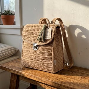 May include: A beige crocheted backpack with a flap closure, a silver metal clasp, and two shoulder straps. A green tassel hangs from the top, and a tag reads THE URBAN CANVAS BACKPACK. A small white label on the side features the text MUSA.